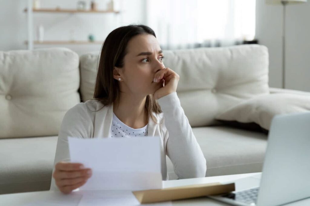 woman sits by couch holding a medical bill and stares into the distance